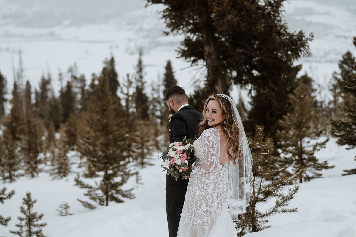 Bride smiles over shoulder while holding bouquet in snowy Colorado forest.