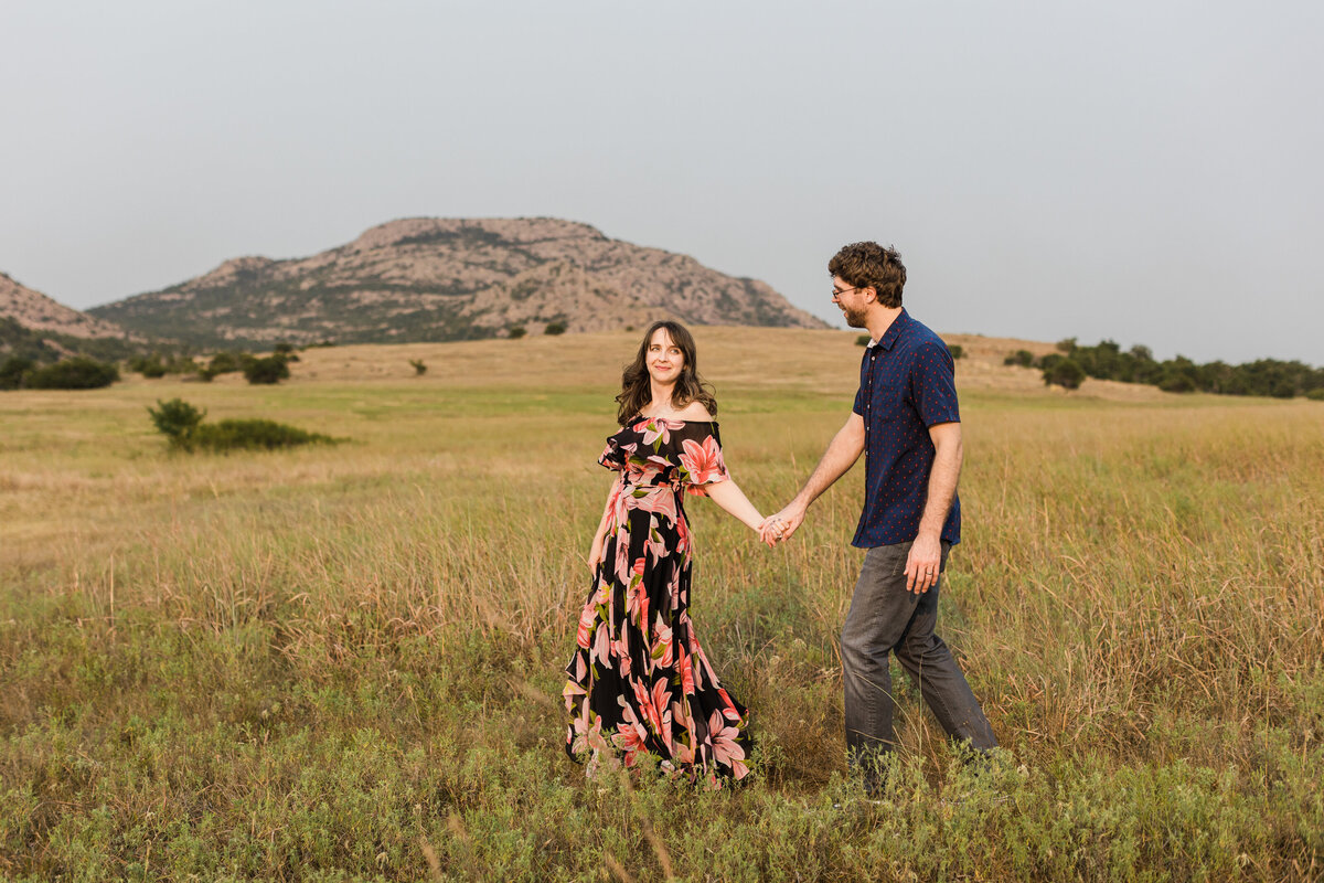 A couple walking through a field during their anniversary session in the Wichita Mountains in Oklahoma. The woman on the left is wearing a long black dress covered in large, colorful flowers. The man on the right is wearing a dark blue dress shirt and dark jeans. A few of the Wichita Mountains can be seen off in the distance.
