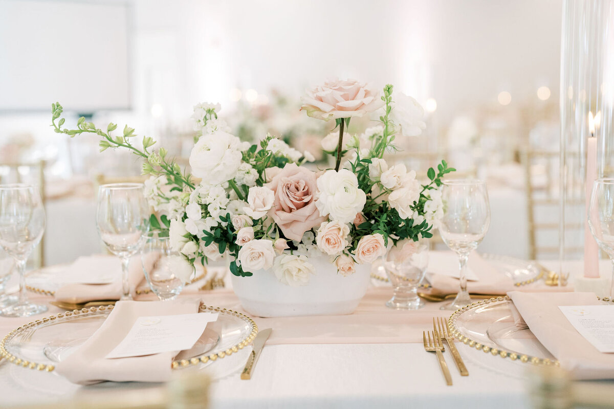 Elegant table setting with a centerpiece of white and pale pink flowers, surrounded by glassware, gold cutlery, and subtle place settings on a crisp white tablecloth at a modern &amp; romantic wedding in Calgary.