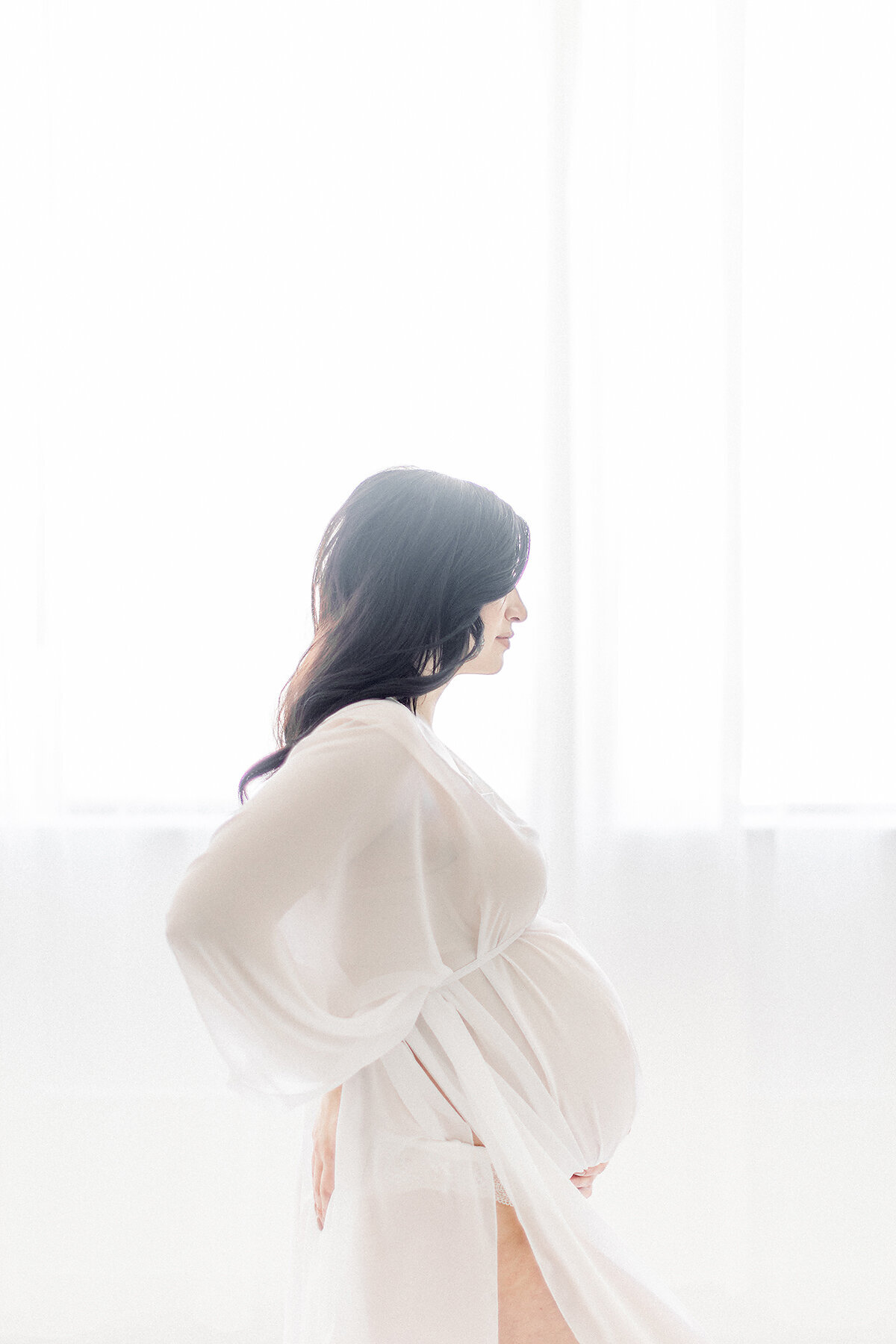 A light filled pregnant mother dressed in a chiffon maternity gown while she is standing in front of a window  at a Dallas/Fort Worth photography studio.