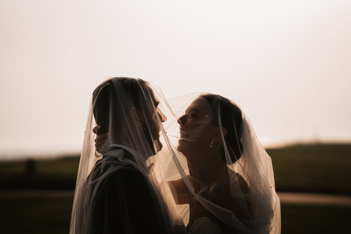 Bride and groom smiling under the bride’s veil during golden hour at a luxury wedding venue in Lytham, capturing an elegant and romantic moment.