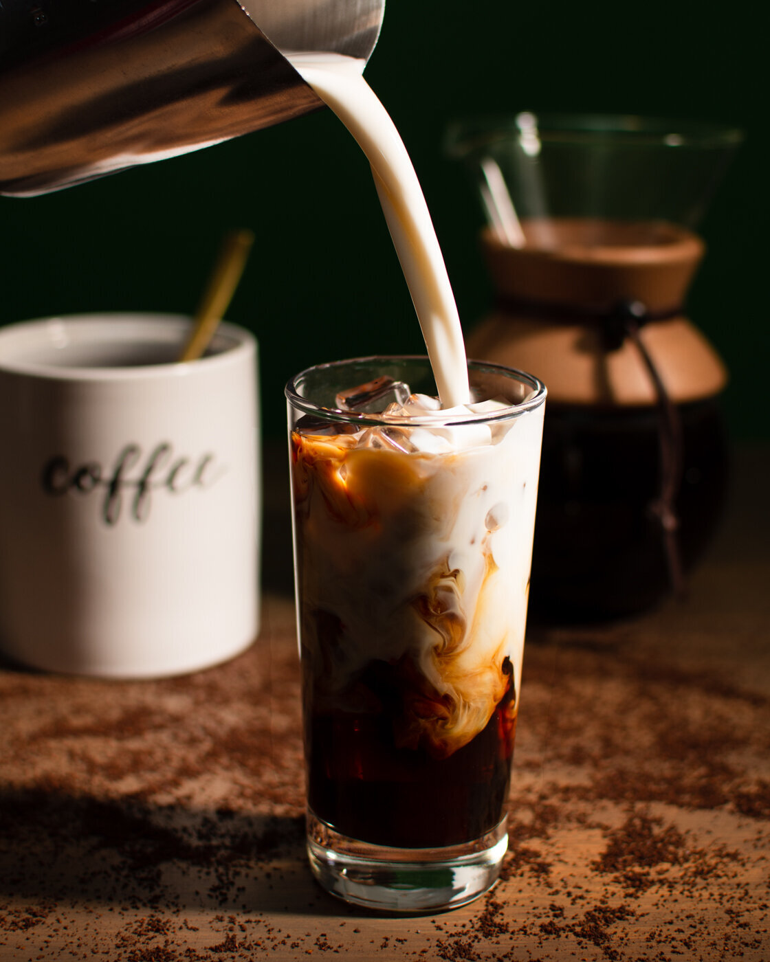 Iced coffee with creamer pouring into a tall glass with a table scattered with coffee grounds and a coffee pour over carafe.