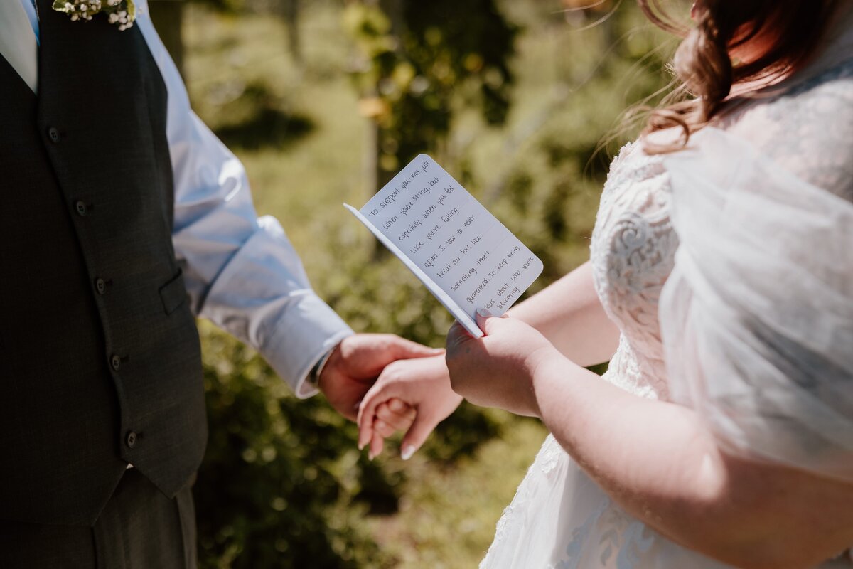 Bride holding groom’s hand while reading handwritten vows during outdoor wedding ceremony.