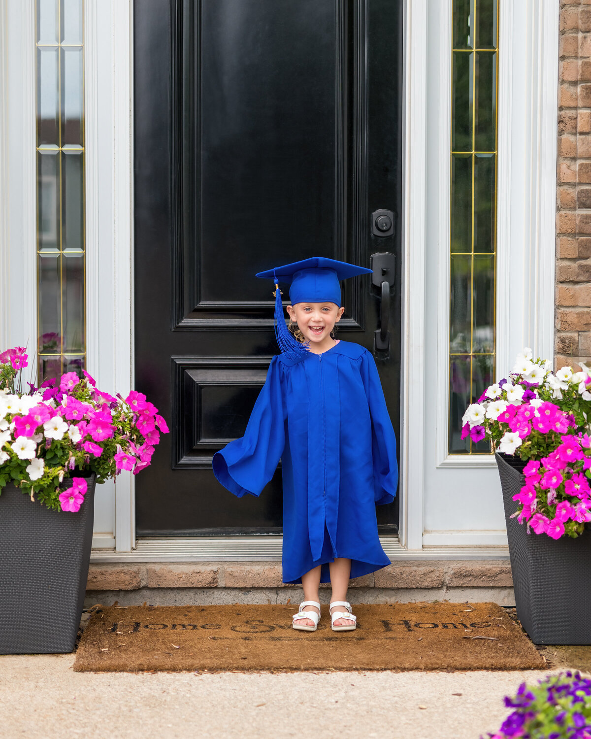 Graduation milestone of a young girl in a blue cap and gown smiling at her front door—beaming with pride and promise.