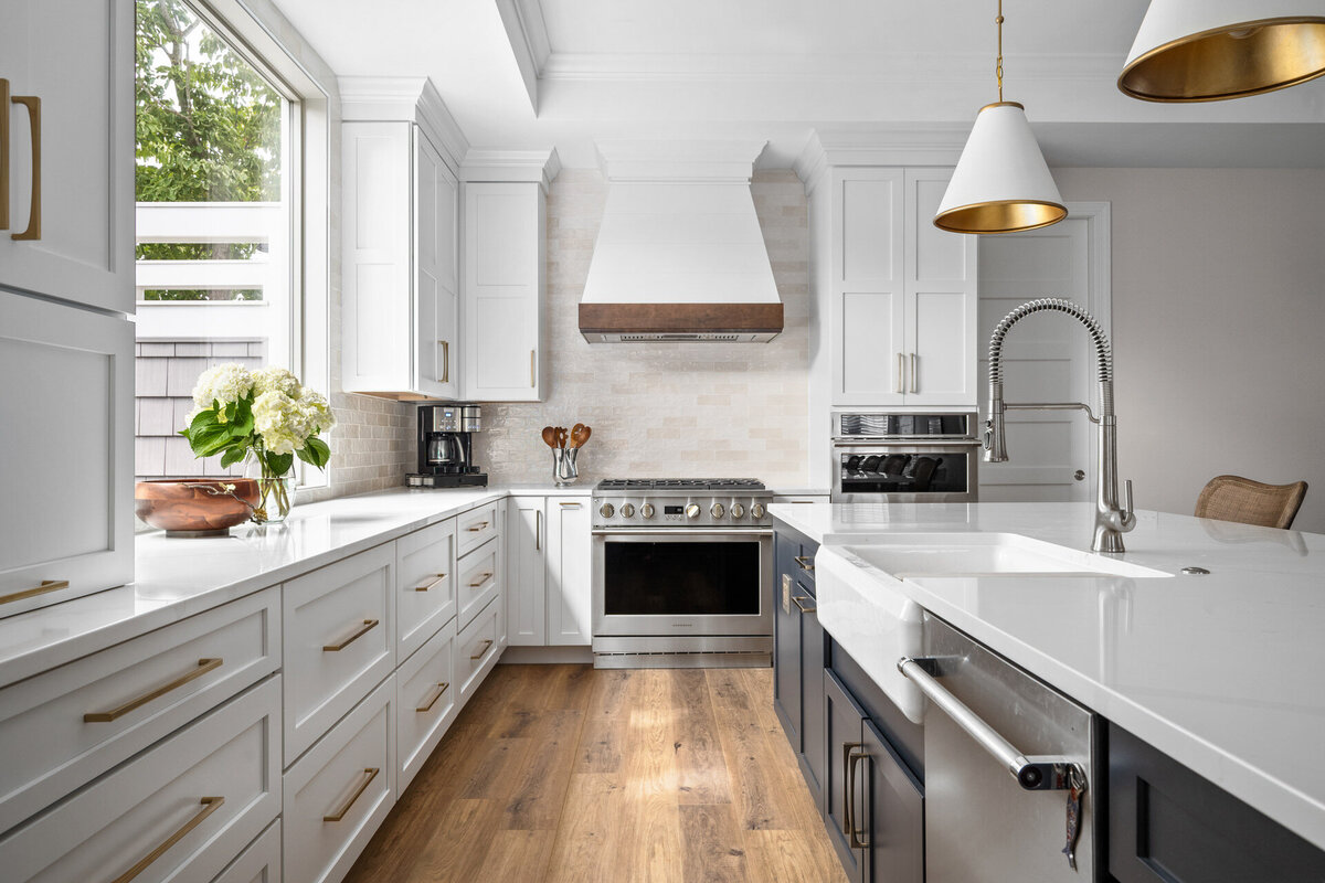 A kitchen in Ocean City, photographed by Svetlana Leahy of Refined Capture, features white cabinets, wood flooring, and a large island. It includes a stainless steel stove with pot filler, elegant pendant lights, and a window view of greenery. The countertops are adorned with flowers and a fruit bowl.