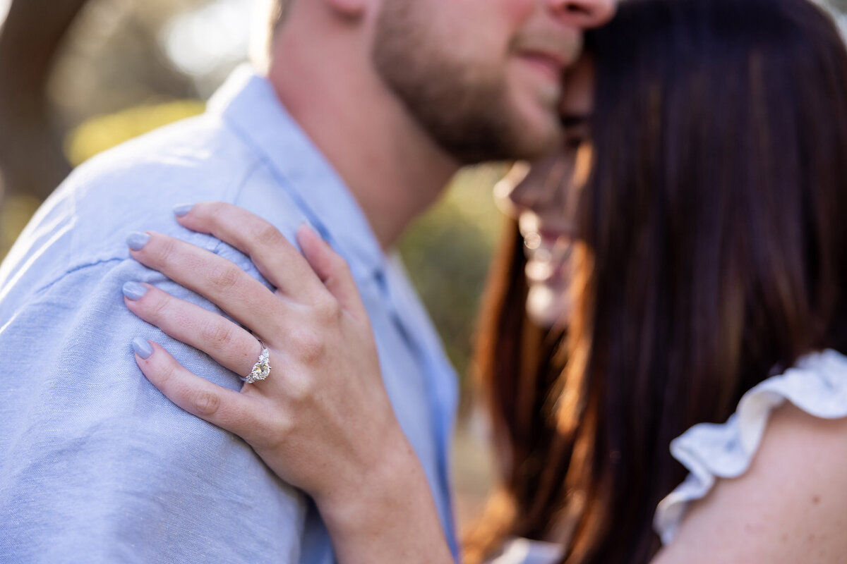 newly engaged couple taking photos at Watercolor park and the picture is of the bride to be's hand in the foreground on the future grooms shoulder and you can see their faces in the background.  Only the ring is in focus by Gwyne Gray Photography