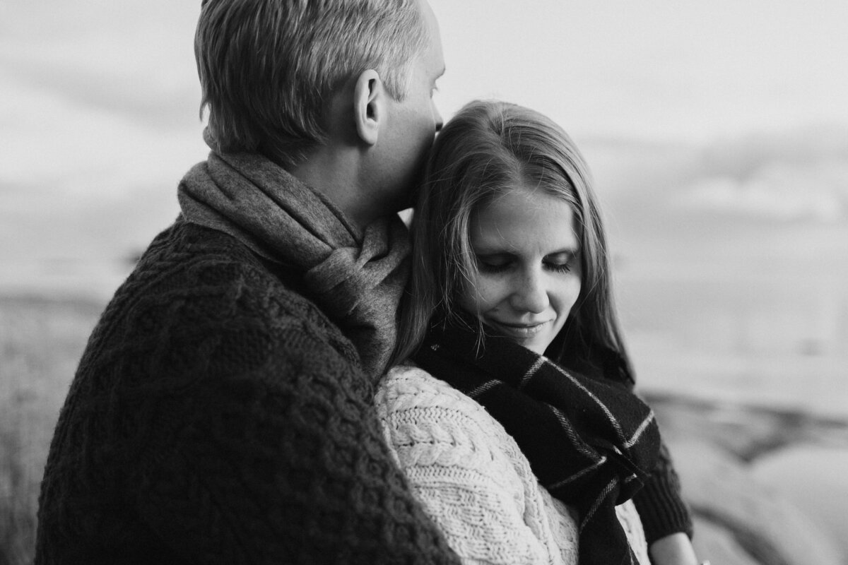 Mies halaa kihlattuaan hänen selkänsä takaa ja nainen pitelee silmiään kiinni ja hymyilee Lauttasaaressa Helsingissä.