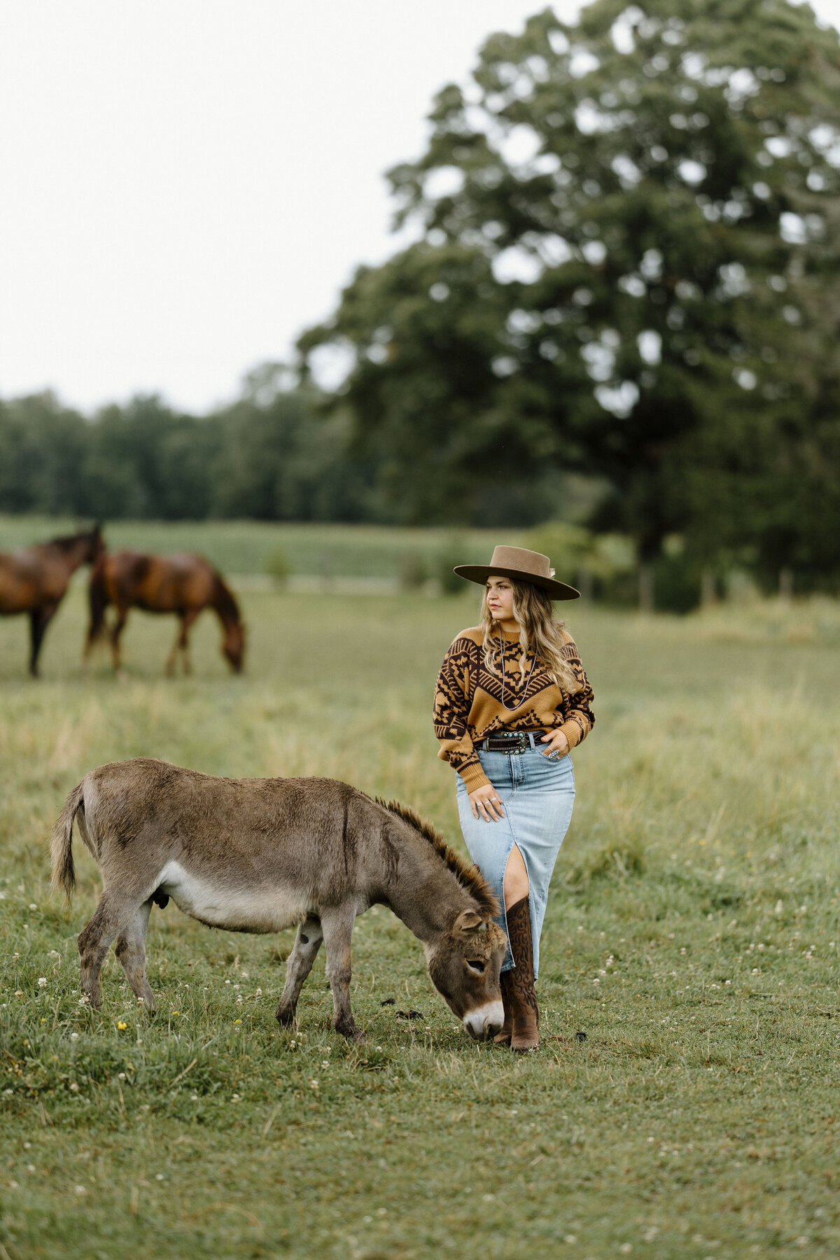 a girl in a pasture dressed in a jean skirt and mustard yellow sweater blouse and cowboy hat standing next to her donkey