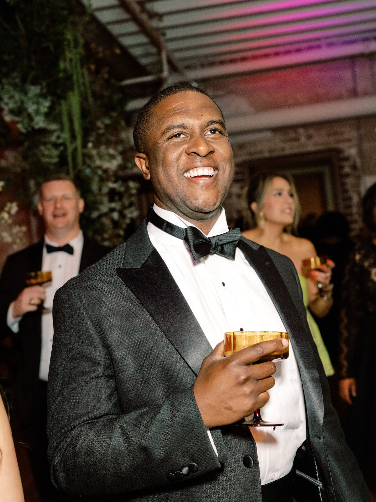 A cheerful man dressed in a formal tuxedo raises a drink in celebration, showcasing a moment of joy and connection among attendees at a lively wedding.