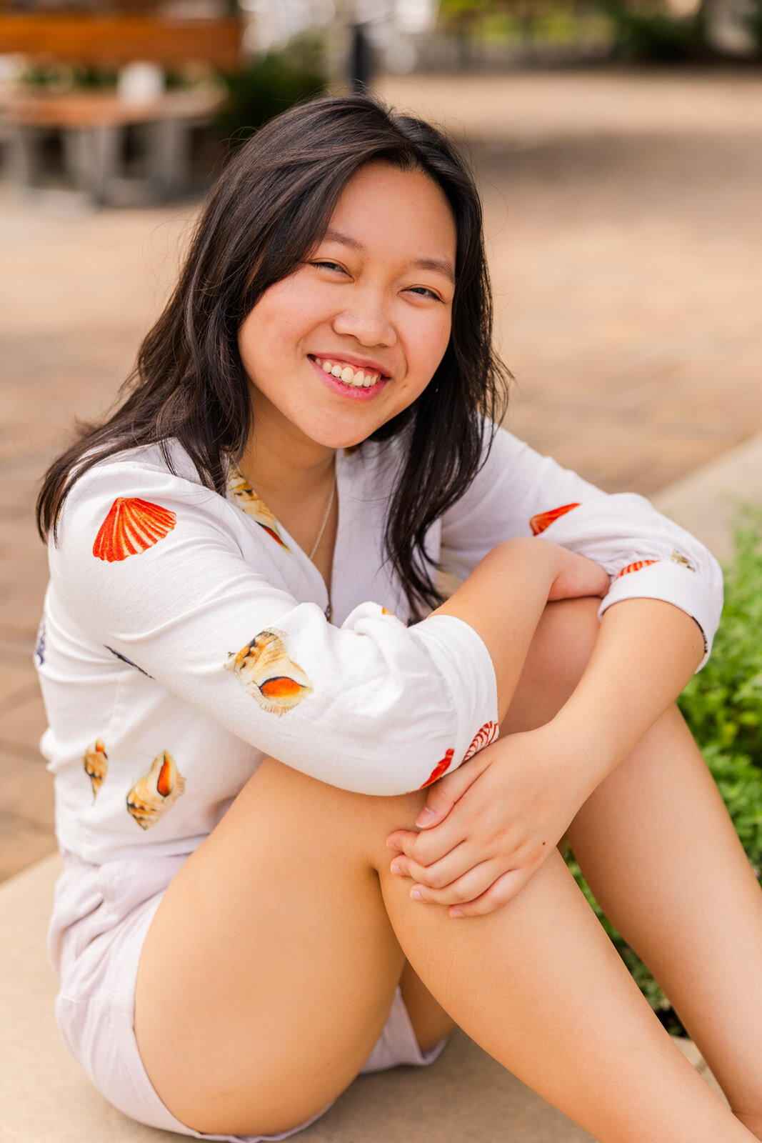 young woman in white shorts and blouse with patterns sitting and hugging her knees during senior session in Sandy Springs GA