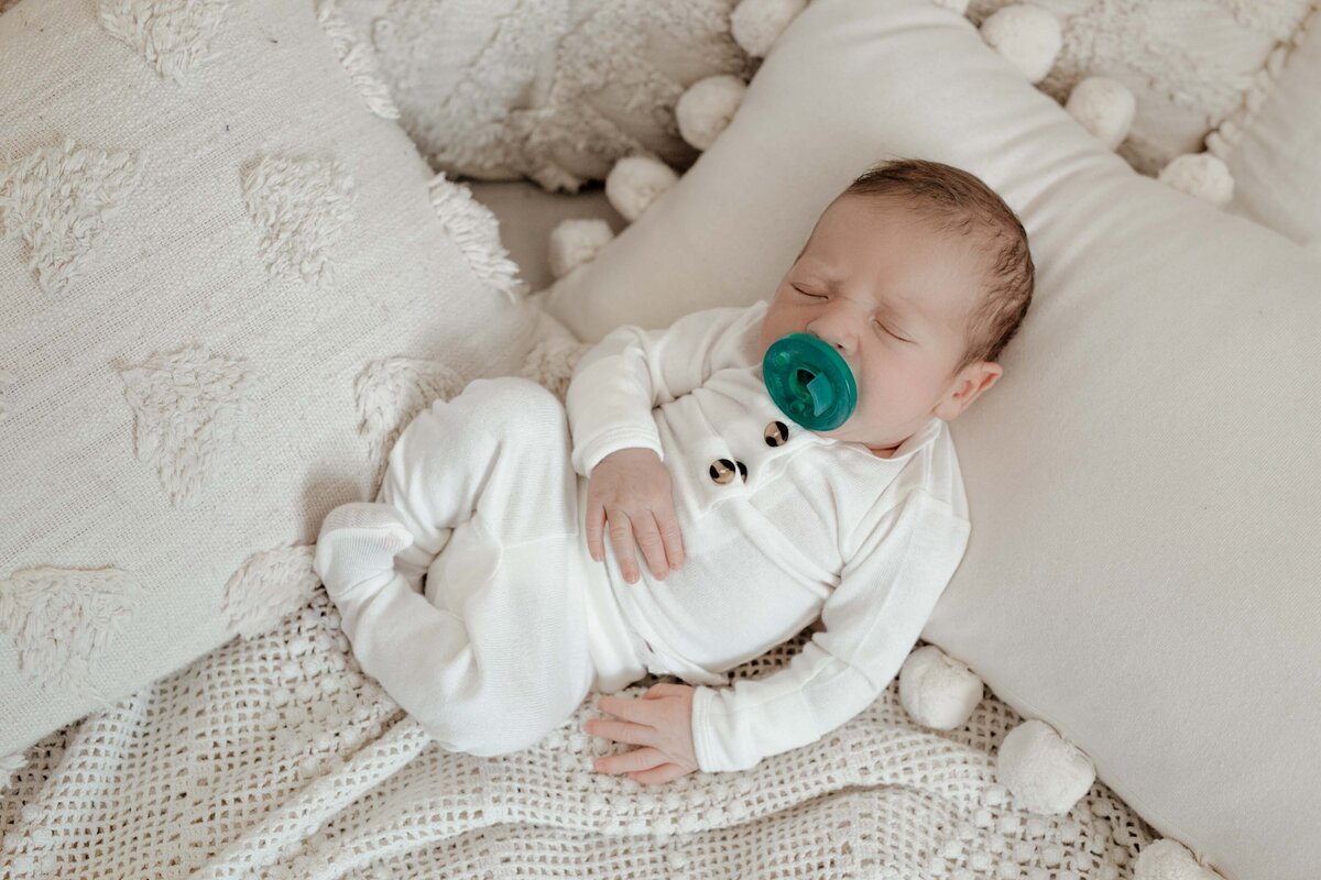 Studio lifestyle newborn session - baby in white onesie sucking on green pacifier laying amongst cream boho blankets and pillows