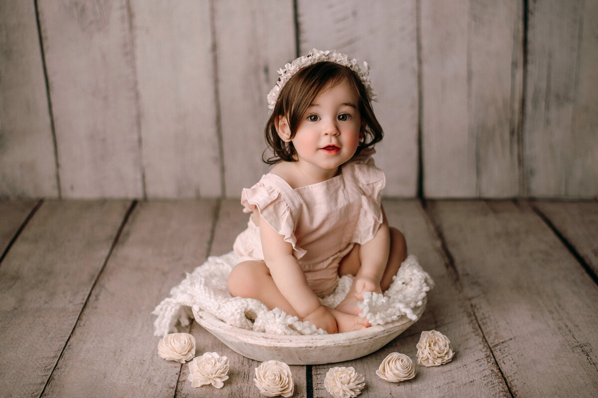 One year old baby girl sitting inside of white bowl prop in front of a cream plank backdrop with flowers on the floor