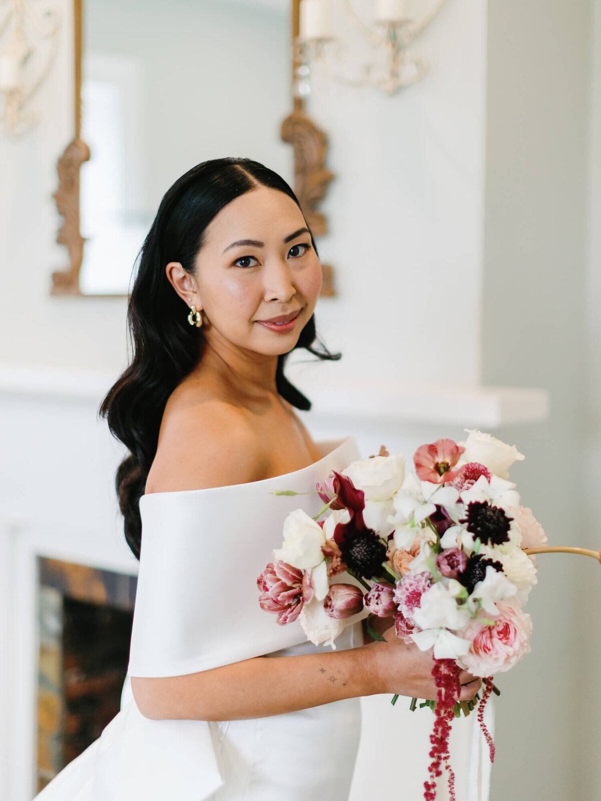 Bride in an elegant white off-shoulder gown holds a bouquet of pink and white flowers, smiling softly.