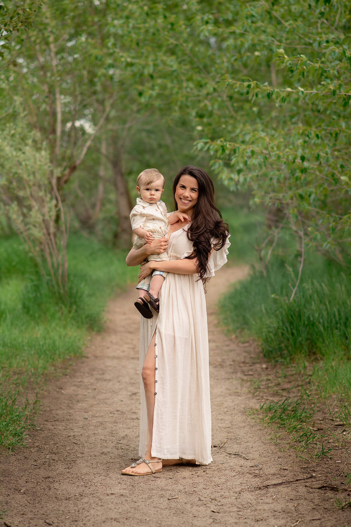 A mom wearaing a long flowy whites dress hold her baby boy on her hip. She smiles and he looks at the camera. Taken in Kamloops by Brittany Danielle
