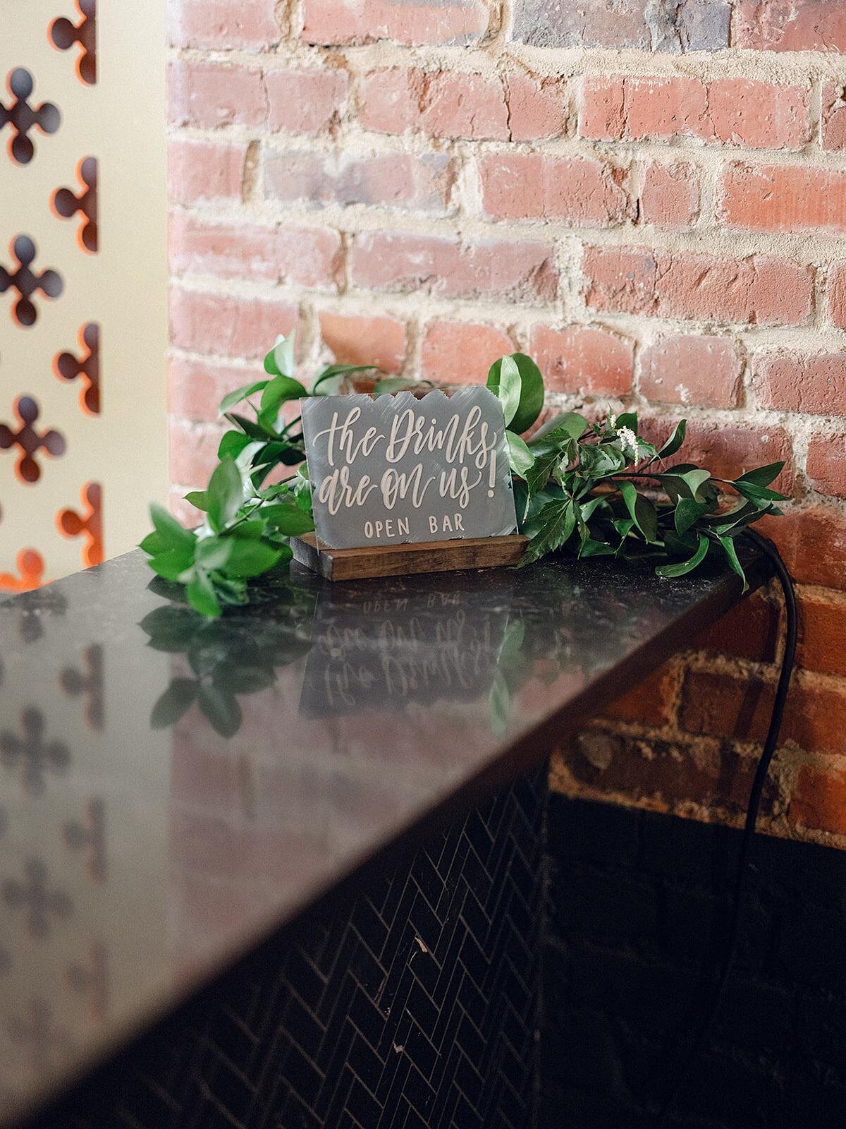 Black granite bar at Clementine hall with an exposed brick wall and gold filigree accents with a  wood bar sign decorated with greenery
