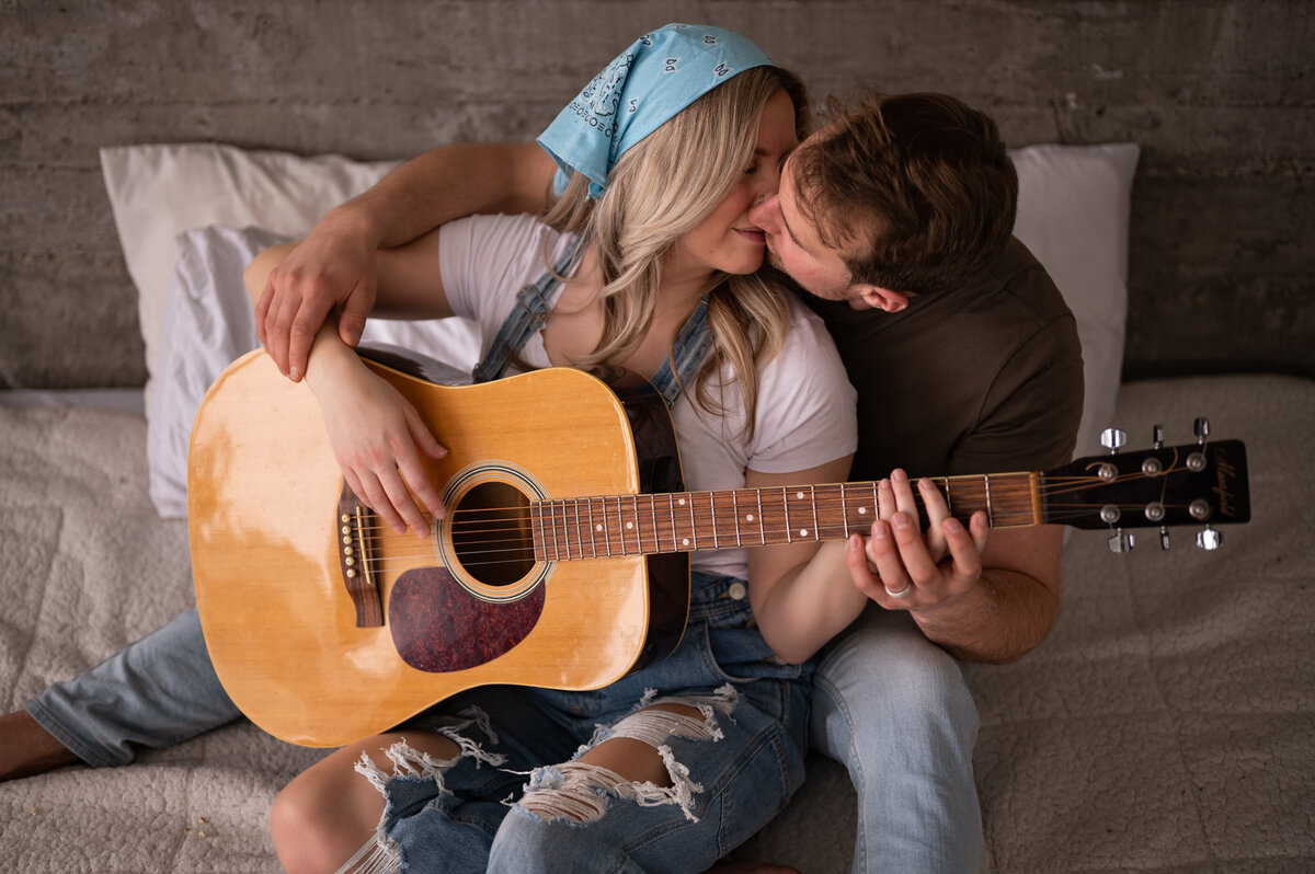 man kissing his wife as he teaches her to play guitar during their engagement photography session at the Pendennis Building in Edmonton, Alberta