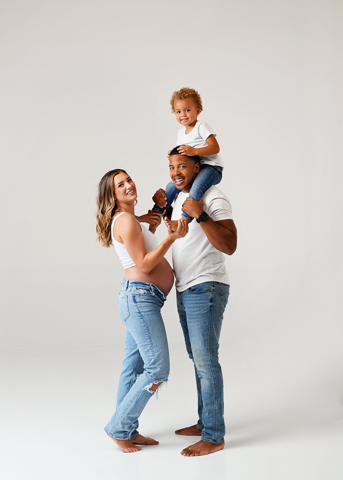 Smiling mom and dad, with toddler on dad's shoulders looking at the camera wearing white shirts and jeans against plain white background with baby bum showing, in Chandler Maternity studio