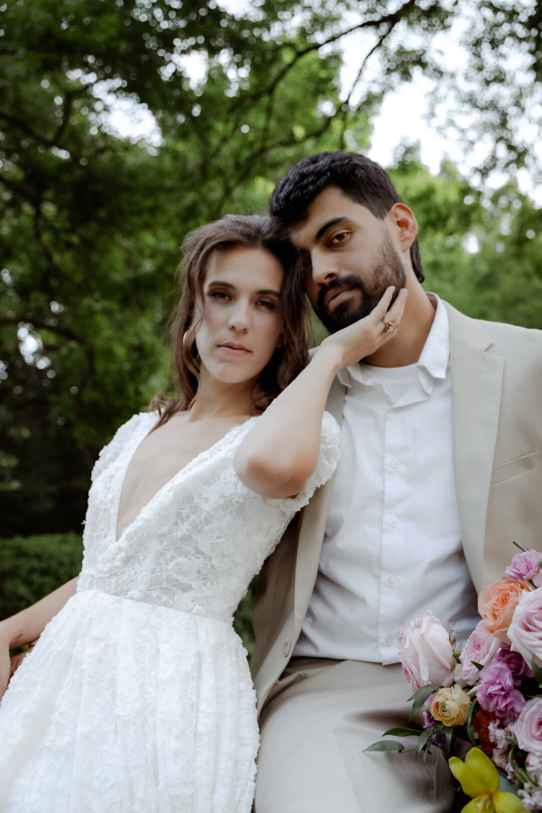 Groom sitting on a tree branch while bride rests her head on his shoulder, holding a vibrant bouquet.