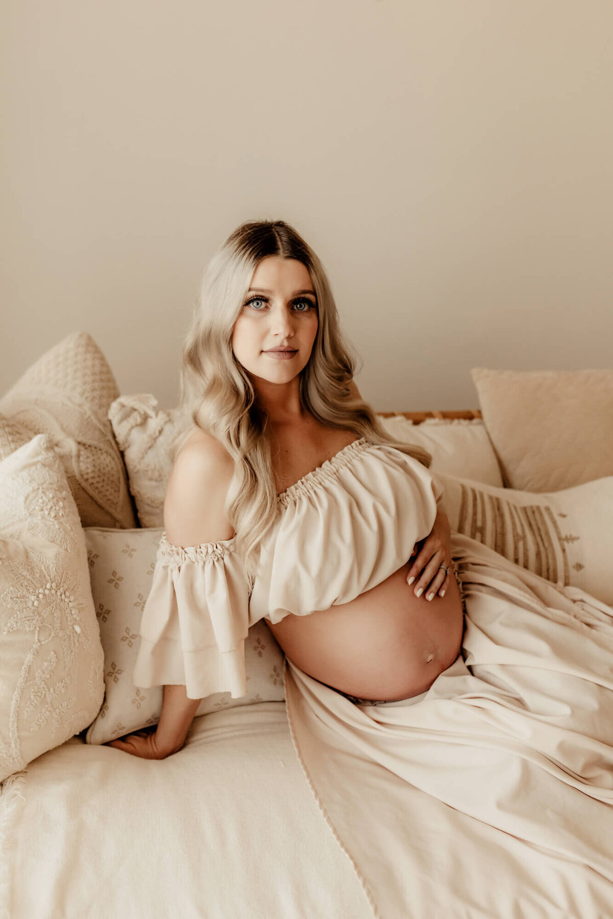 Mother sits on a daybed and looks at the camera for a maternity portrait.
