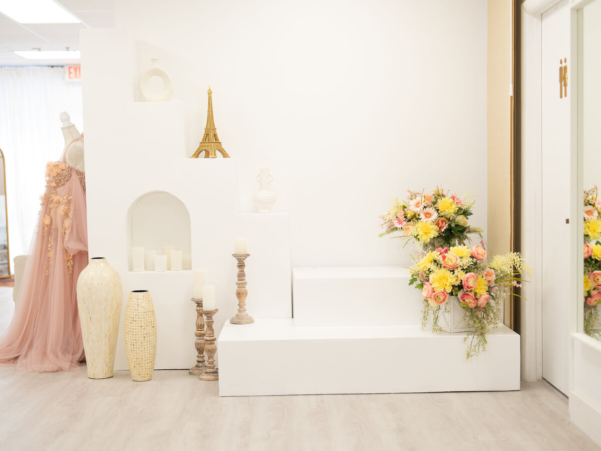 Spacious room with a white display featuring candles, a vase, and an Eiffel Tower model. A pink gown and vibrant flower arrangement add elegance.