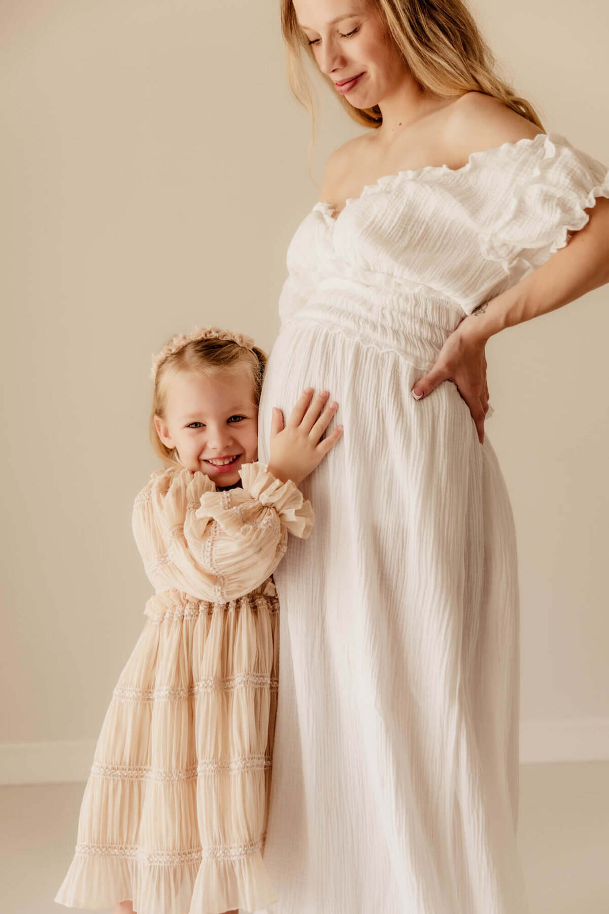 Girl wearing a pink dress smiles while holding her mother's baby bump.