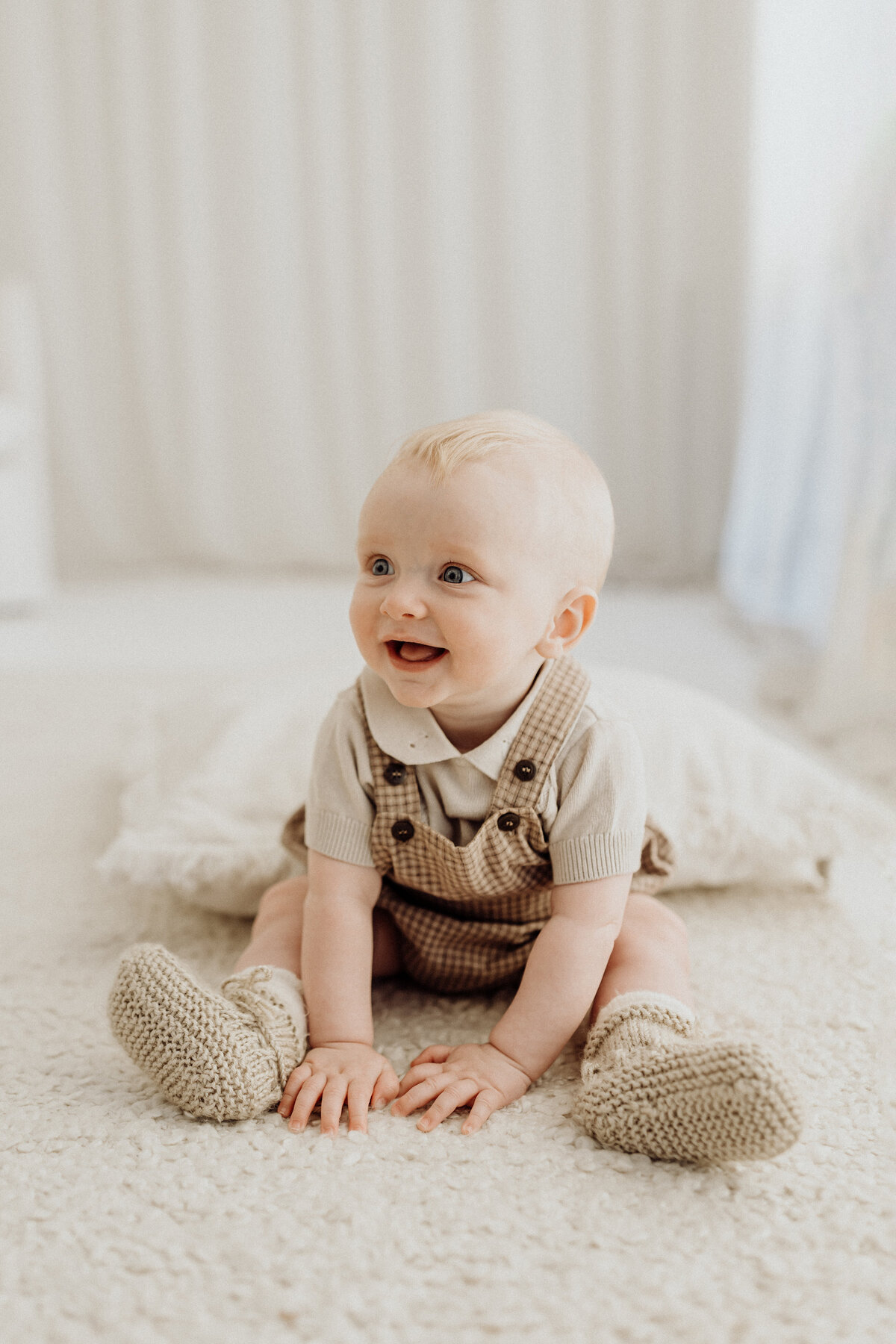 A baby sitting up with his little booties on and a checkered set of overalls, next to the window in the studio while he giggles and lucy jane photography takes his milestone portraits in christchurch