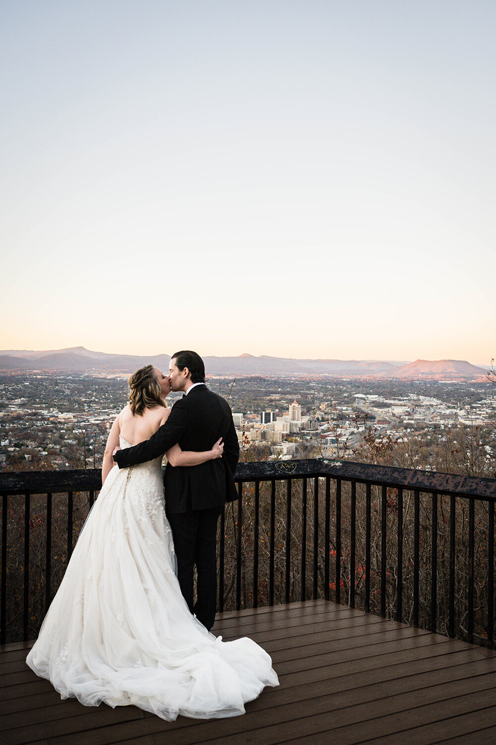Two marriers on their elopement day face the cityscape of Downtown Roanoke and the Blue Ridge Mountains at sunset at the Rockledge Overlook in Virginia. The couple wraps their arms around the other’s back and goes in for a kiss.
