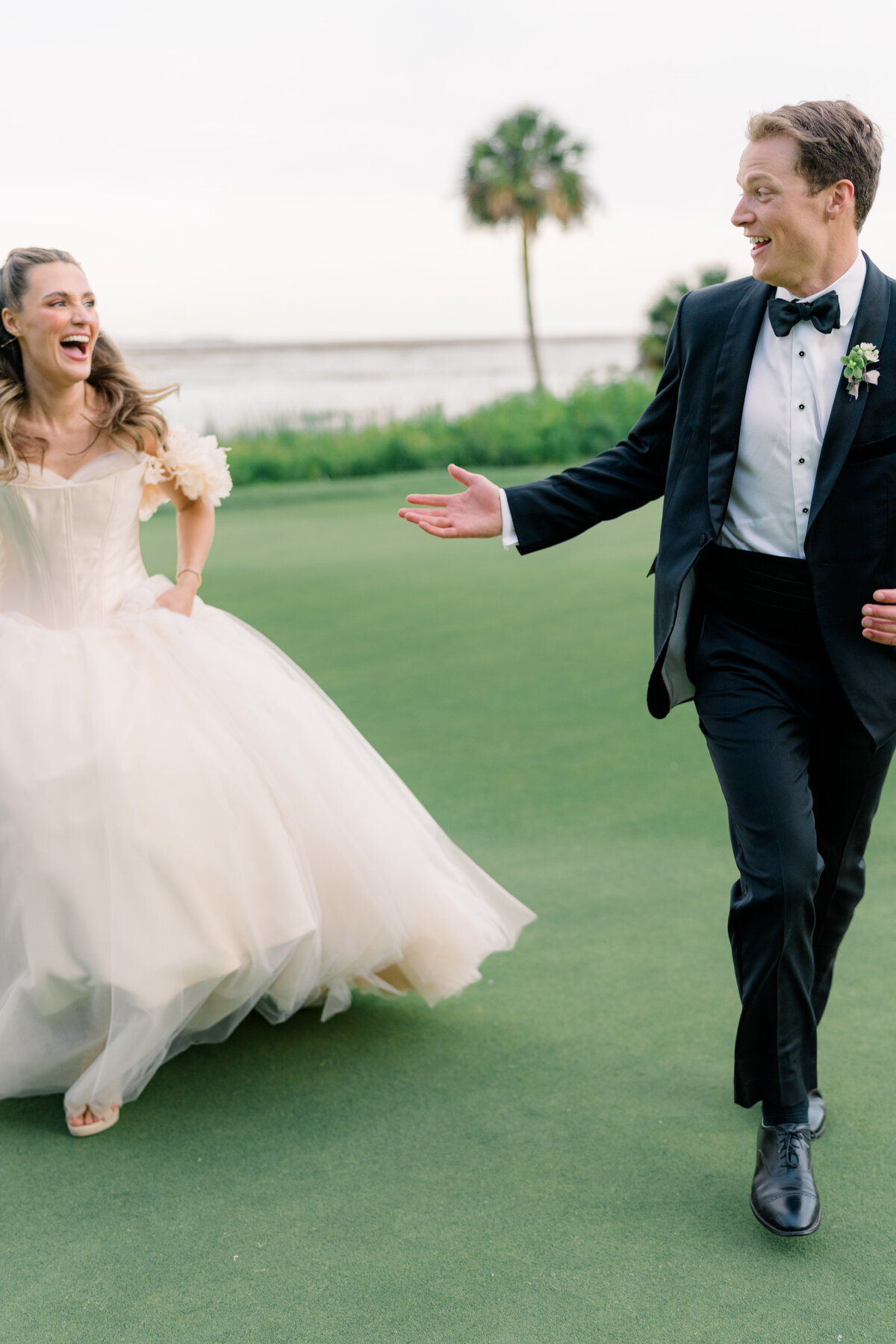 Bride and groom running together with palm tree in the background. Blush wedding dress with floral sleeves, corset top, and long tulle bottom. Spring Island Destination Wedding. Old Tabby Links. Southeast Destination wedding photographer
