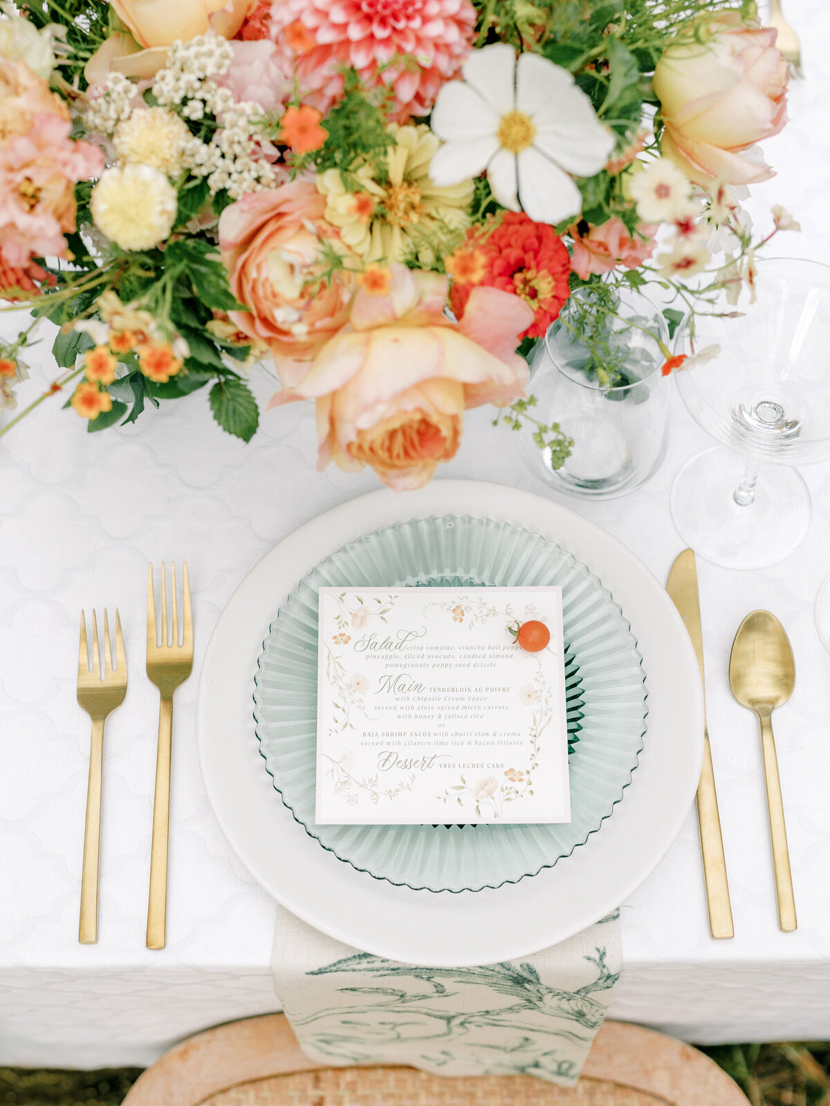 Top down fine art photography of a table setting with a white charger, green plate, gold serving-ware with a menu on top of the plate