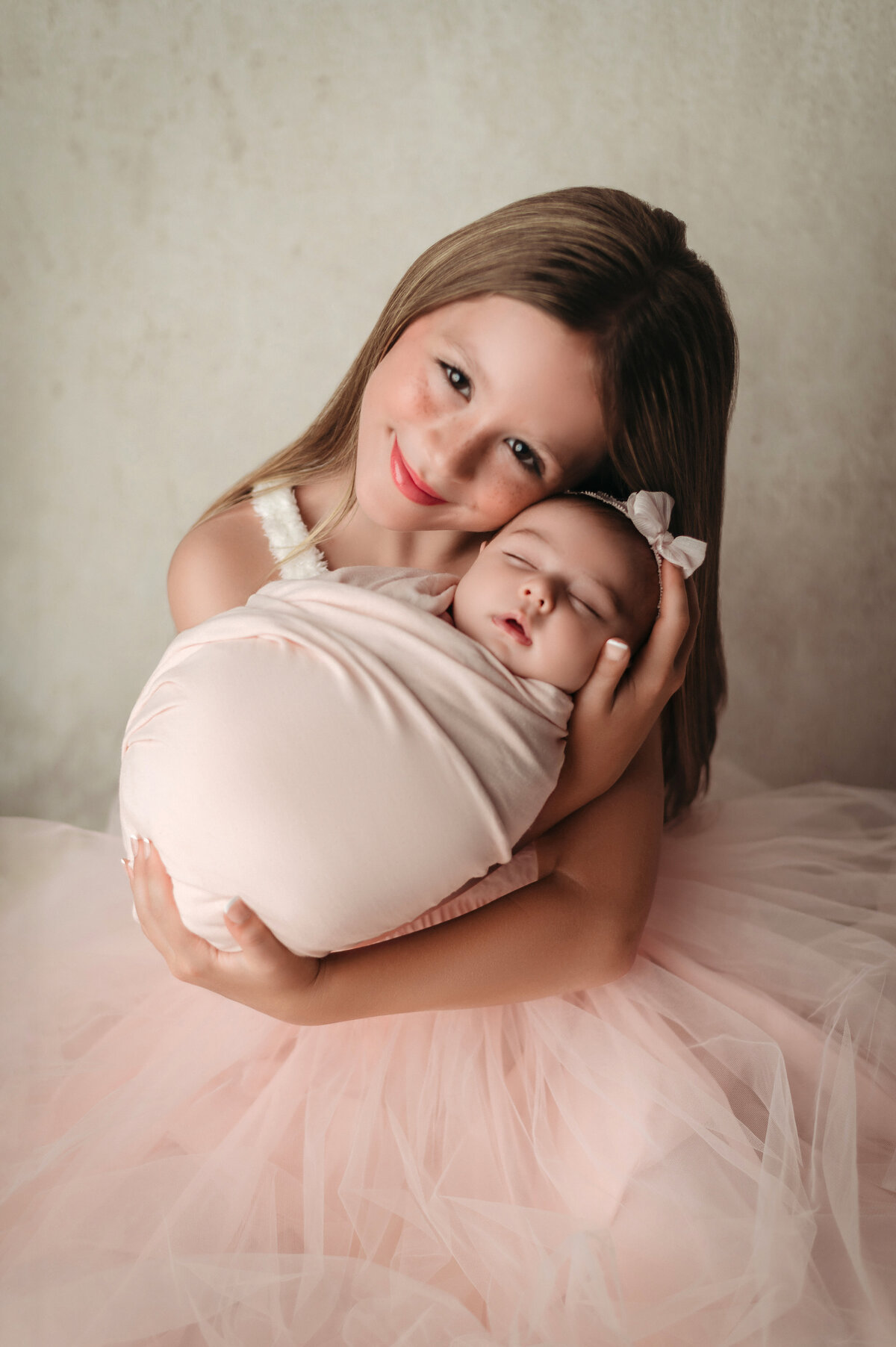 Big sister wearing pink tutu dress holding her swaddled and sleeping new baby sister in her arms and snuggles her