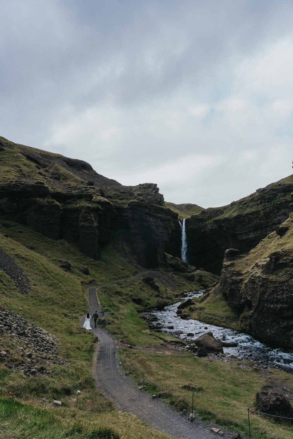 The couple strolls along a quiet trail, hand-in-hand, near a waterfall in Iceland 