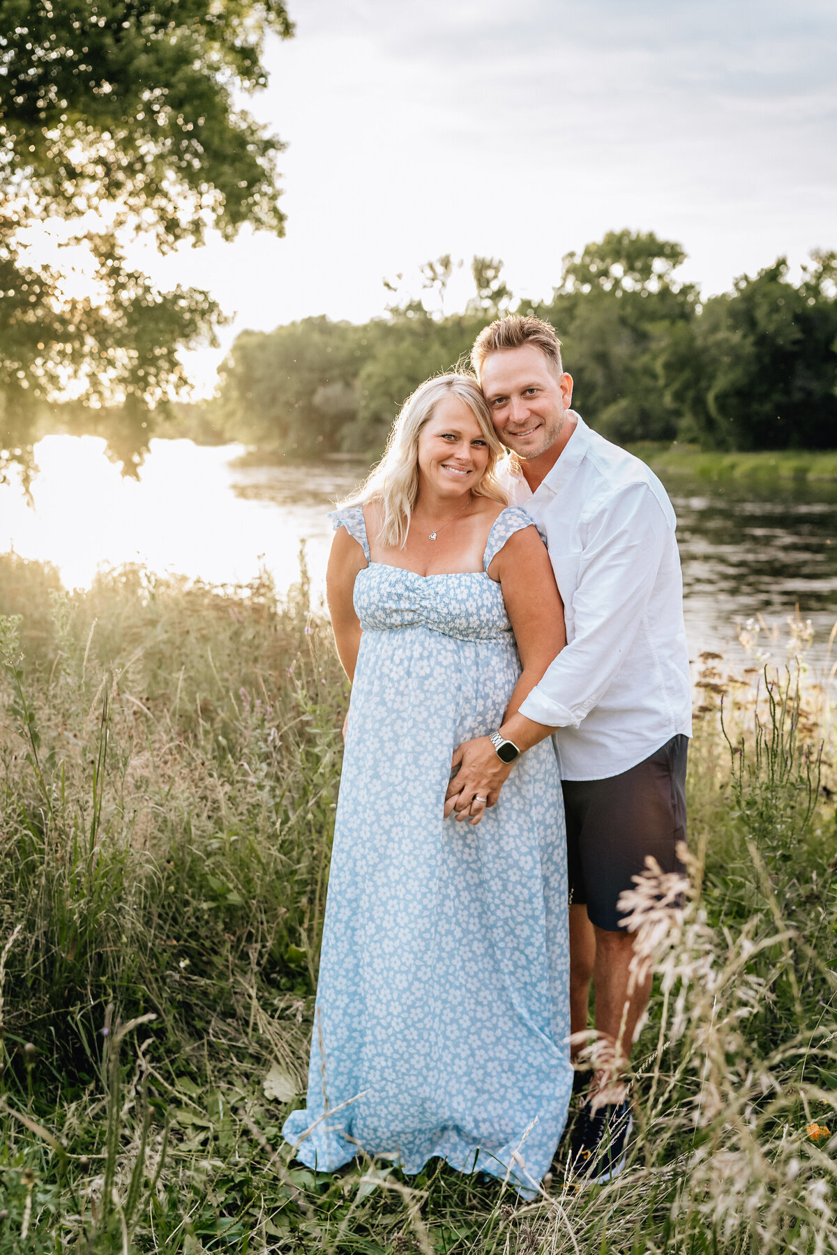 The couple stands in a grassy area by the river, smiling at the camera. The woman, in a light blue floral dress, is pregnant and cradles her belly, while the man, in a white shirt, stands closely behind her with his arms wrapped around her.