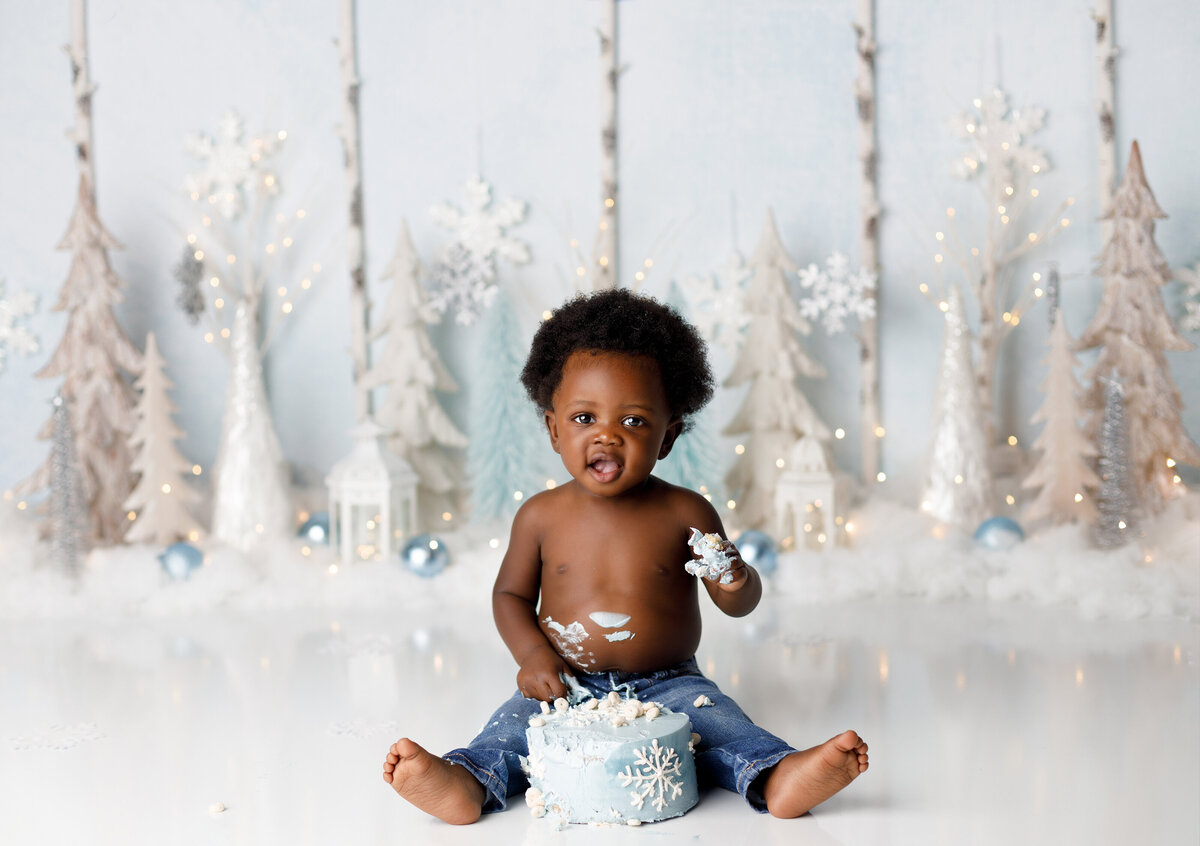 Winter Wonderland cake smash in top newborn and cake smash photography studio in West Palm Beach and Delray Beach, FL. Black baby boy is wearing only jeans with a blue and white cake with snowflakes in front of him. He has icing on his hand and is smiling at the camera. In the background are white trees, faux snow, and snowflakes.