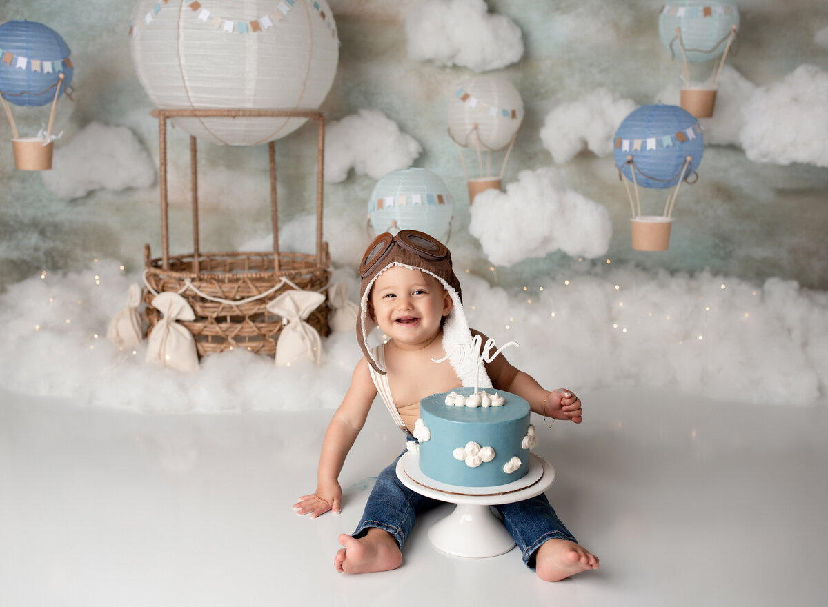 Baby boy hot air balloon themed cake smash with West Palm Beach and Boca Raton cake smash photographer. Baby boy wearing jeans and vintage pilot hat with blue iced cake with clouds on it between his legs. He has a big smile and looking at the camera. In the background, there are 5 hot air balloons of various sizes in shades of white and blue.