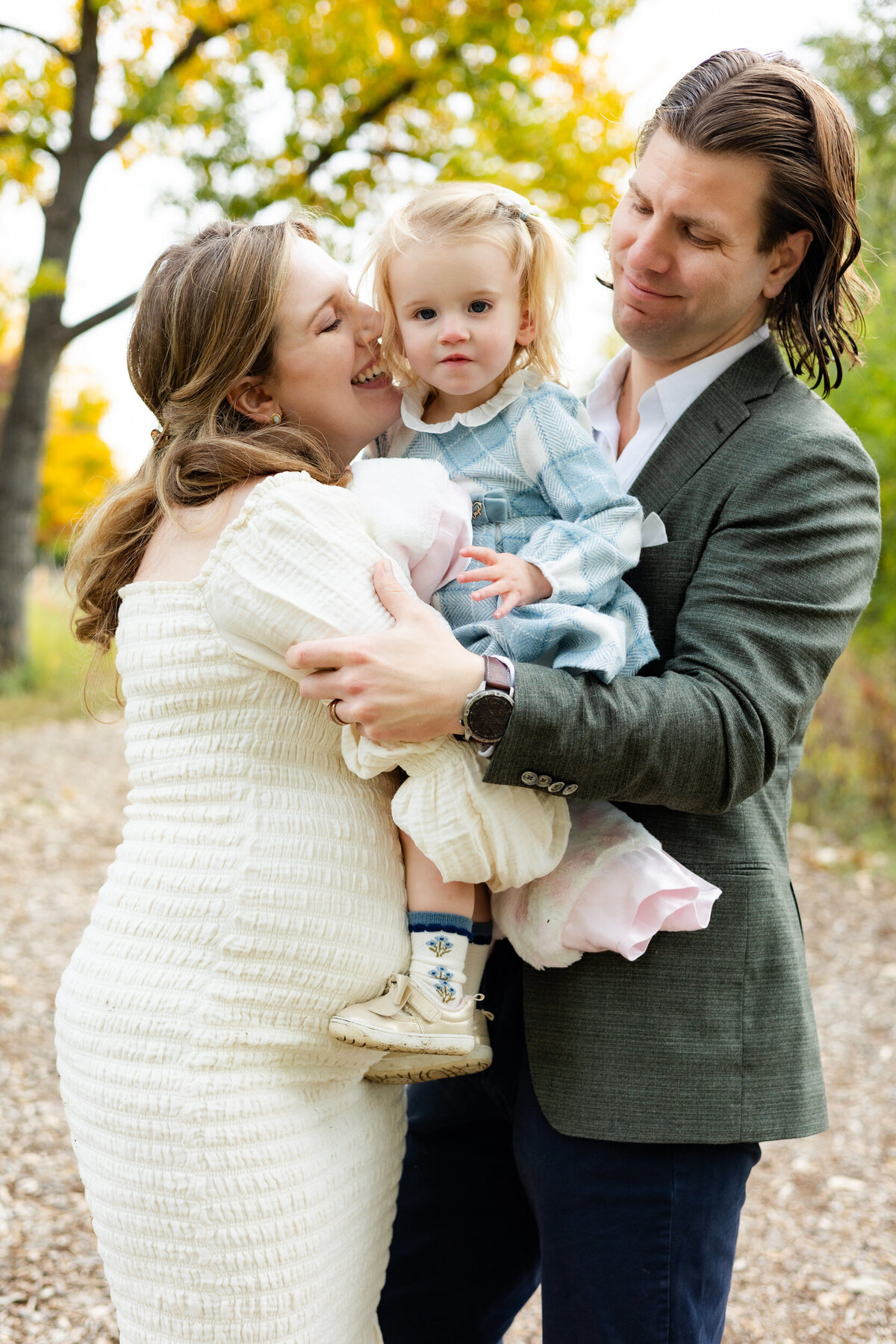 Mom and dad hold toddler daughter between them and snuggle and smile at her while she looks at the camera.