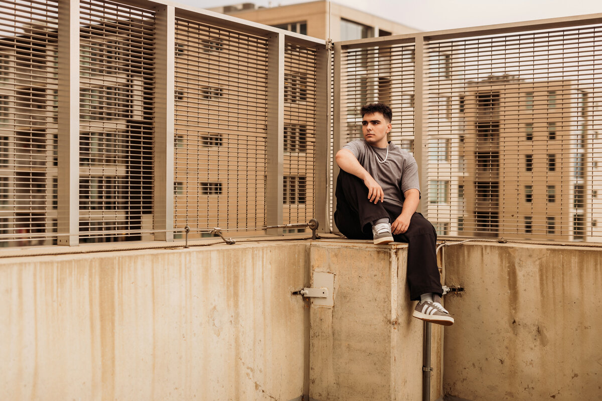 high school senior boy sits on top of concrete pillar with metal gates behind him watching the city skyline for his urban senior photos