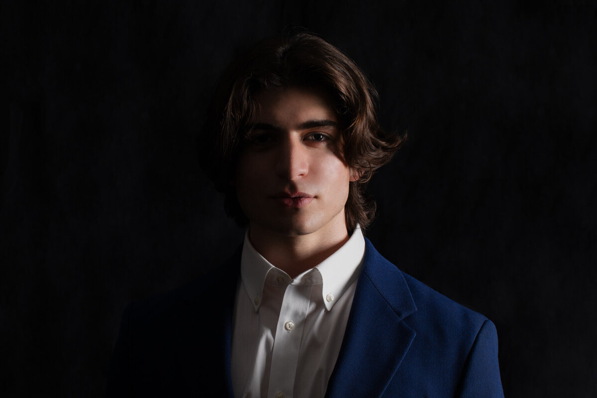 A close-up of a young male model with black hair wearing a white dress shirt and a navy suit jacket, standing in the dark with half of his face shadowed.