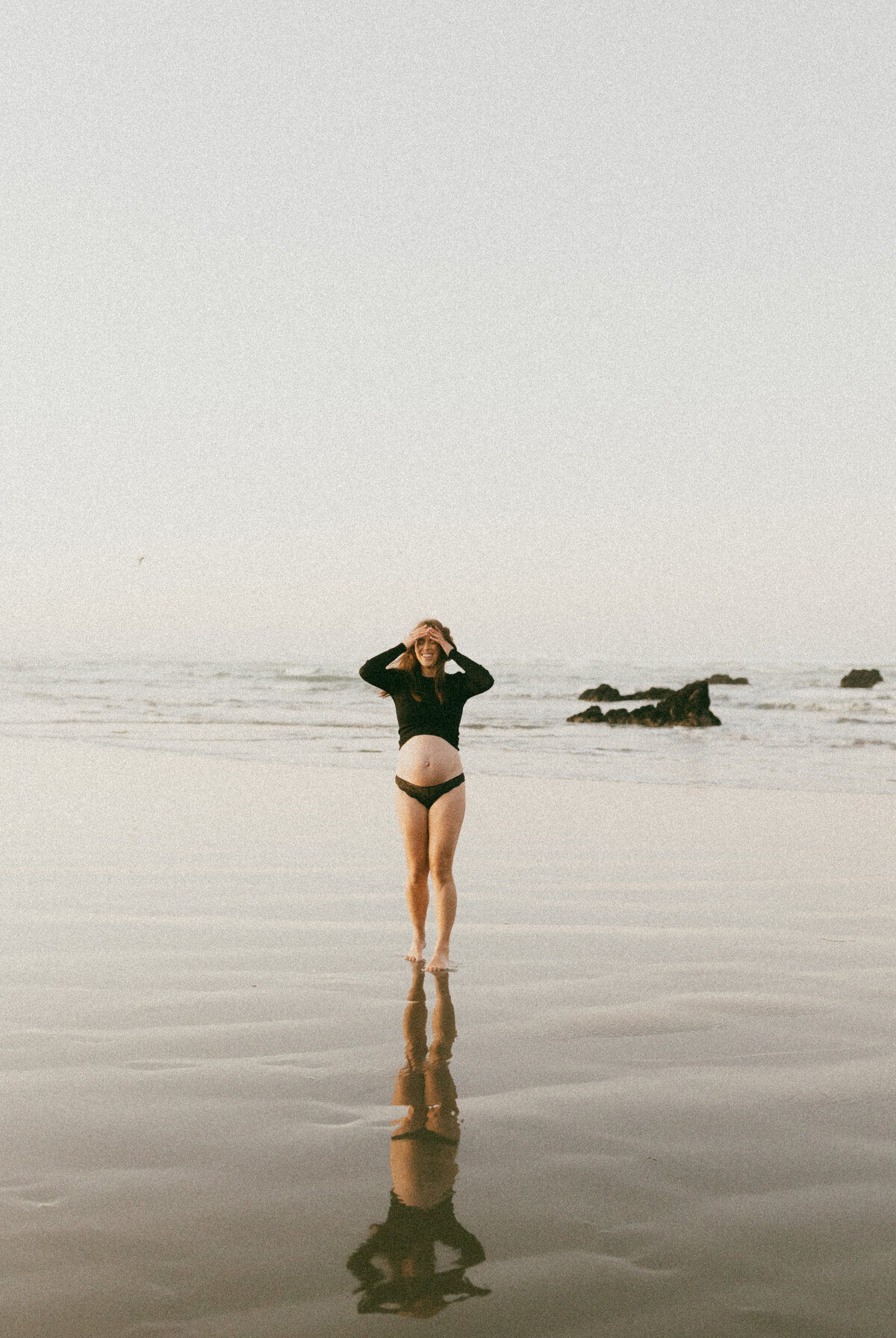 A woman wearing a black top walks along a beach with her hands in her hair during a maternity photoshoot with christchurch photographer lucy jane