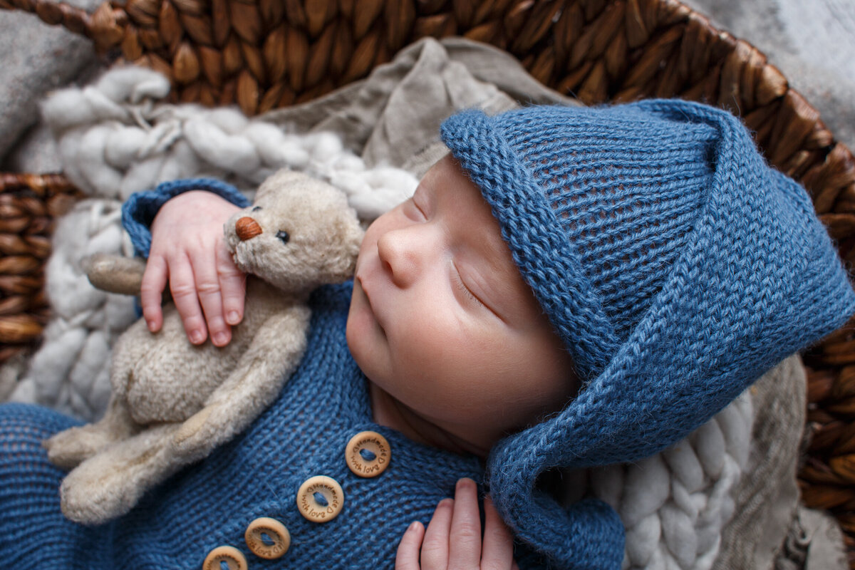 Cose up of Tiny infant wearing a blue sleeper and cap holding a  little stuffed teddy bear