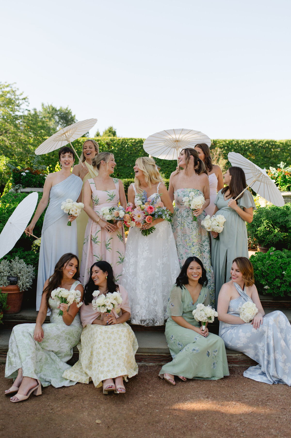 Bridesmaids with the bride, holding parasols and bouquets in an elegant garden setting.
