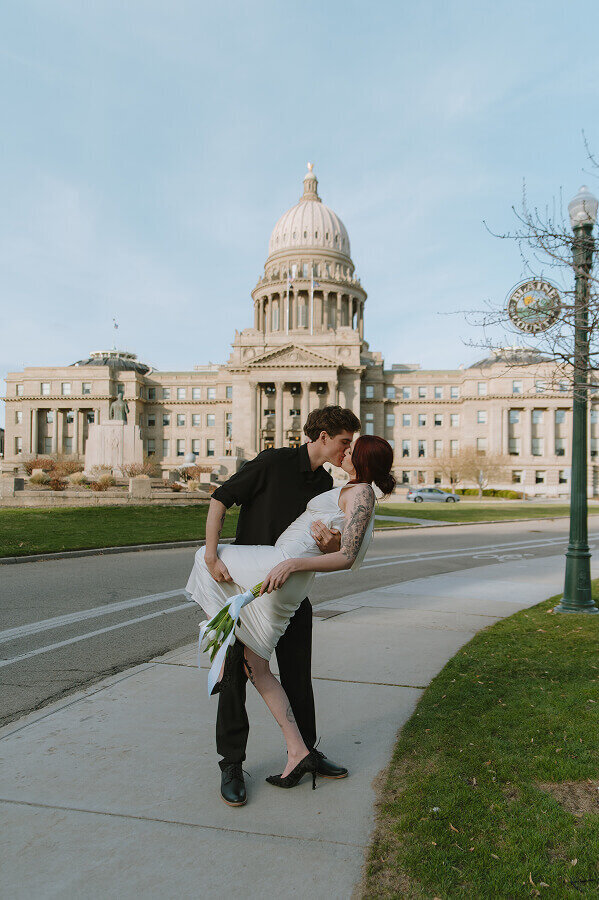 A man slightly dips his bride on in front of the Boise capitol building. He's kissing her and holding up her leg.