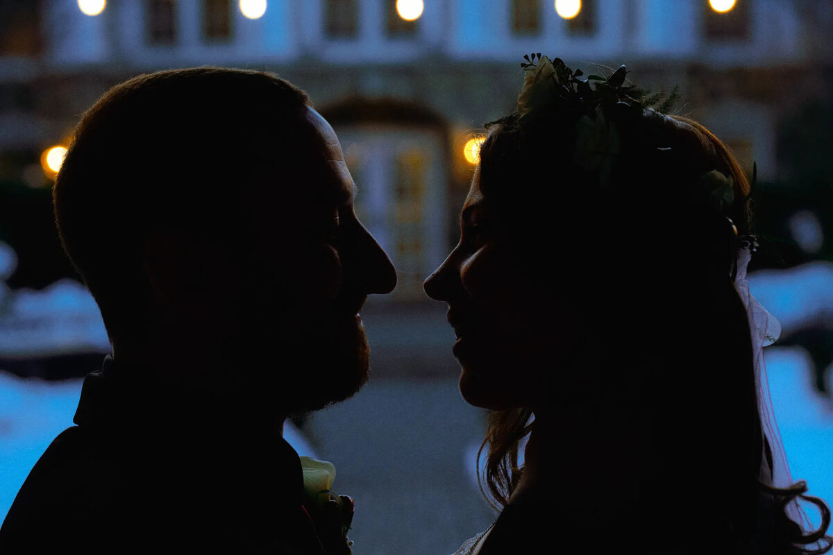 Silhouettes of a man and woman facing each other closely, possibly at a wedding, with soft lights and a blurred building in the background. Captured by an NJ wedding photographer, the scene feels intimate and romantic.