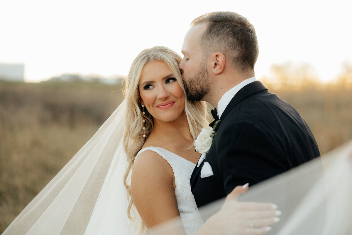 The groom kissing the bride's temple as the veil is draped around them.