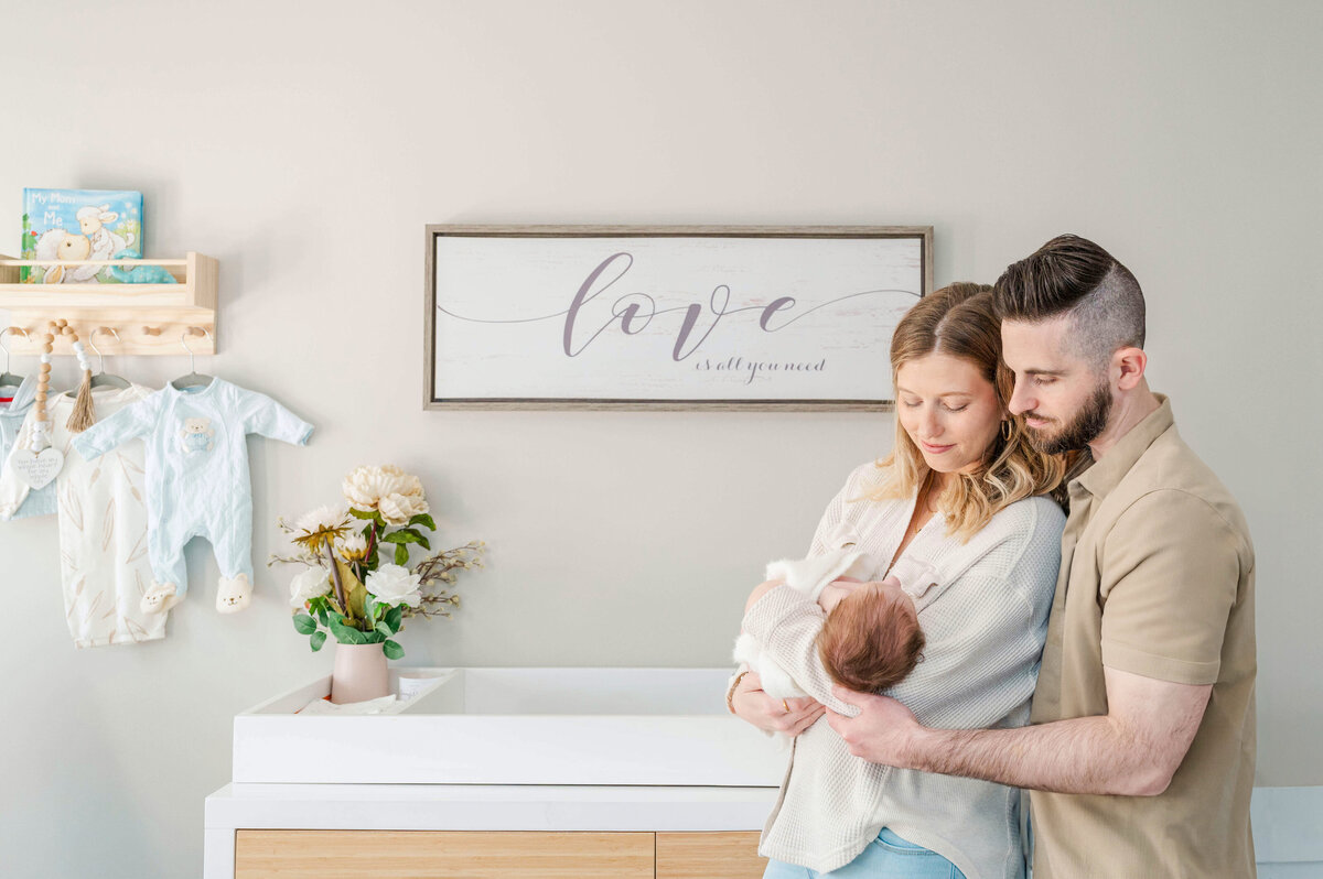 Parents cradeling their newborn and looking down at the baby in front of a changing table in the nursery.