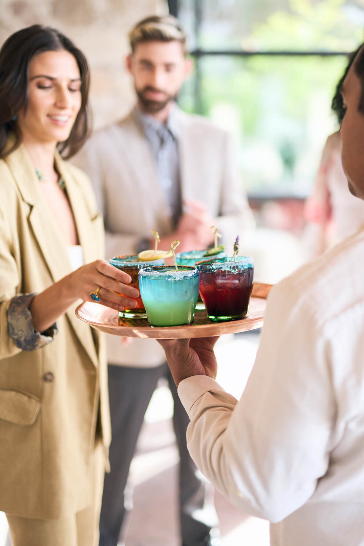 A person offers a tray with colorful cocktails to a woman in a beige suit. A man in a grey suit stands in the background, holding his own drink. The setting appears to be a social event with bright natural lighting.