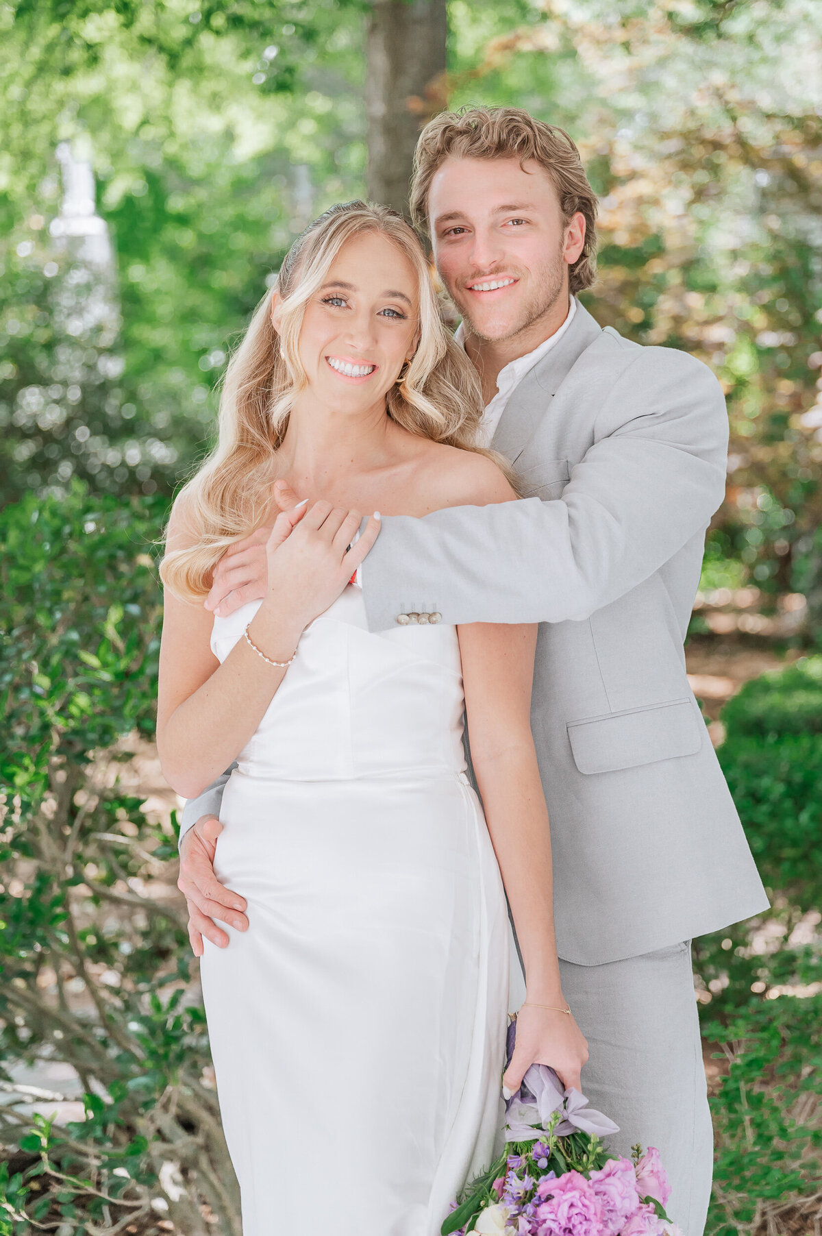A blonde bride holding a pastel purple wedding bouquet being hugged by her husband in a grey suit at The Leslie Alford Mims House enjoying their Raleigh wedding photos by JoLynn Photography