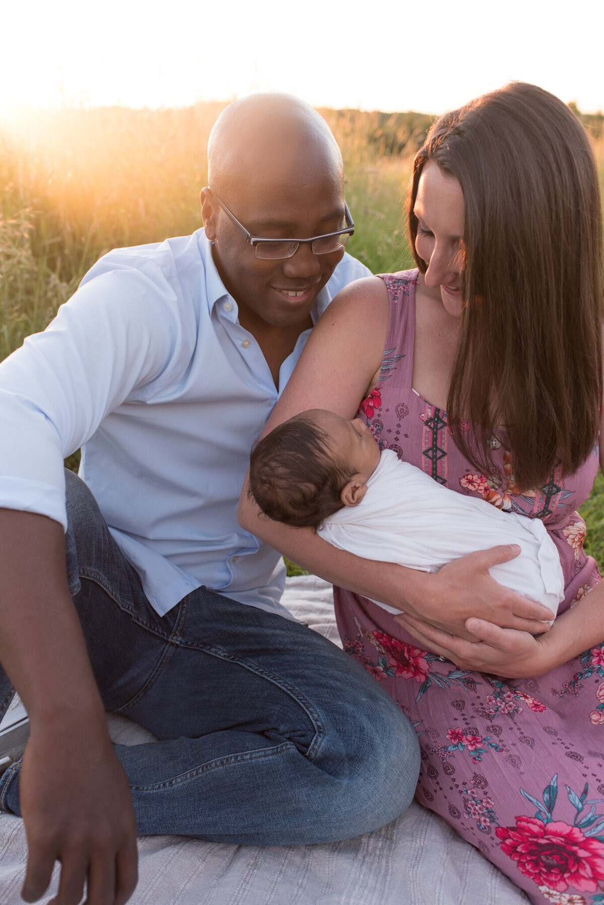 New parents are holding their newborn at an outdoor newborn session in a field with the sun setting behind them