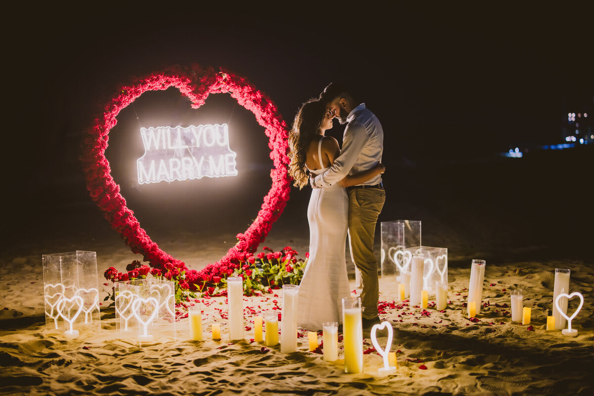 A couple sharing a moment after the proposal in front of an led sign and floral heart arch and romantic candle lighting at long branch beach nj