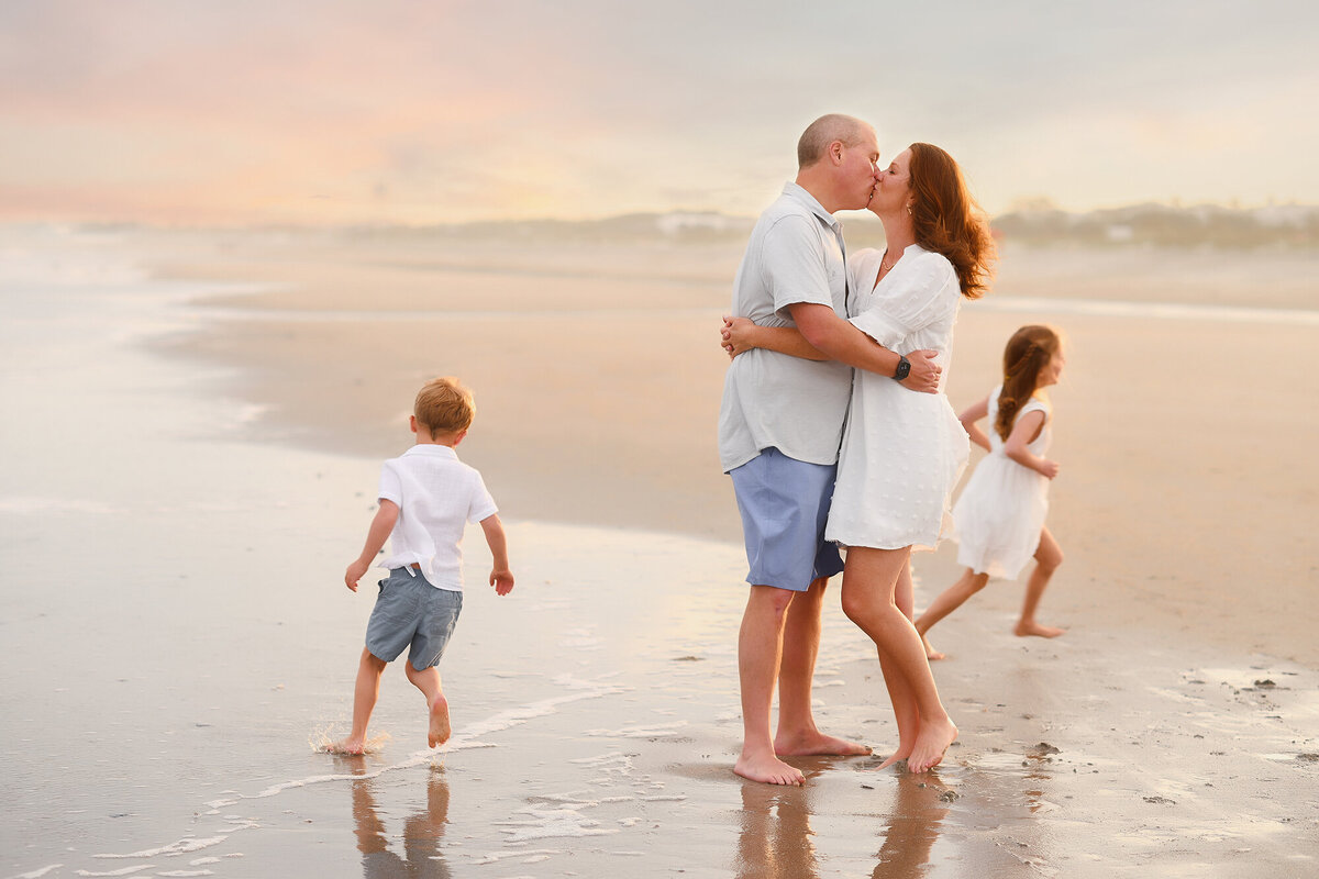 Parents share a kiss while their children run circles around them  during Family Pictures on Isle of Palms. 
