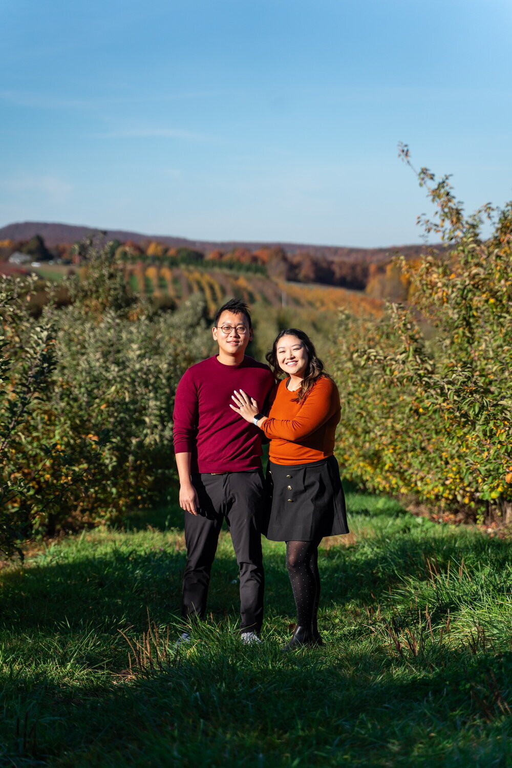straight Asian couple wearing bright red and orange sweaters pose in front of Lyman Apple Orchard in the fall